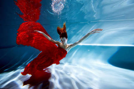 Underwater in the pool with the purest water. Beautiful girl in a scarlet dress and flowing hair.の写真素材