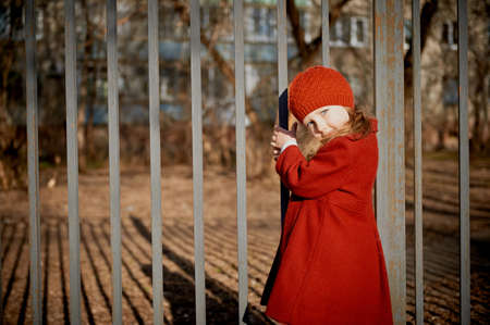 Baby 3 years with long hair. In a red beret and coat stands on the street in the sunshine, near the fenceの写真素材