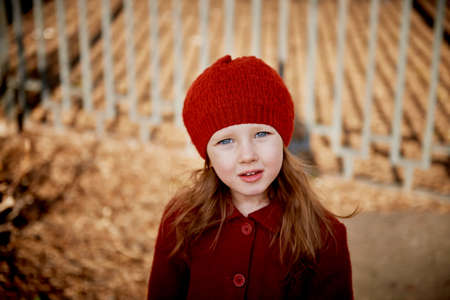 Baby 3 years with long hair. In a red beret and coat stands on the street in the sunshine, near the...