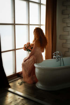 Beautiful red-haired girl romantically sitting on the edge of the bathtub and holding a bouquet. The face is not visible on the background of plants with broad green leaves. In lace dress powdery pastel cream colorの写真素材