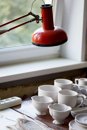 The workplace of ceramist. Baked dishes, ready for painting. A window table with a red lampの写真素材