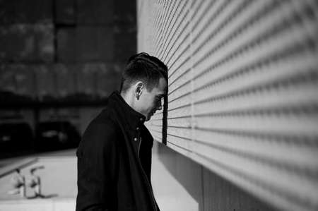 The young man is unhappy in a dead end or depression. Stands leaning his forehead against the wall. Black and white pictureの写真素材