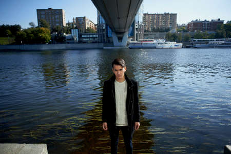 A young man, thin. With dark hair and brown eyes. Standing under the bridge at the river, look at the camera. People in the big city. Lifestyleの写真素材