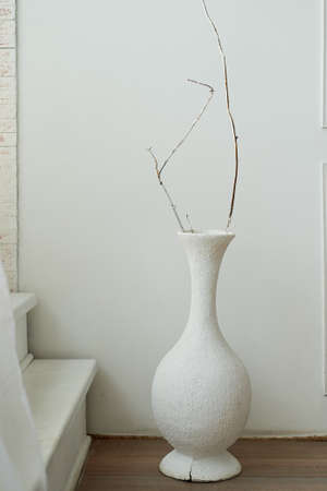 White vase with dry branch stands in front of a white wall. The minimalistic design of the roomの写真素材