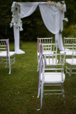 Wedding ceremony in the open air. Transparent chairs with fluttering ribbons in the Park or forest.Arch decorated with white chiffon and flowersの写真素材