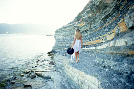 A beautiful girl in a white tunic walks along the Bay of the sea, rocky shore. In the hands of bears black a wide-brimmed hat. Freedom and serenityの写真素材