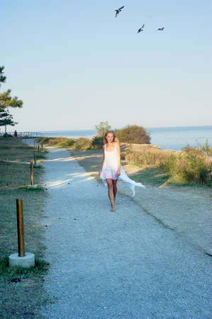 Happy girl in a white tunic is on the path near the sea, the sky flying birds.の写真素材