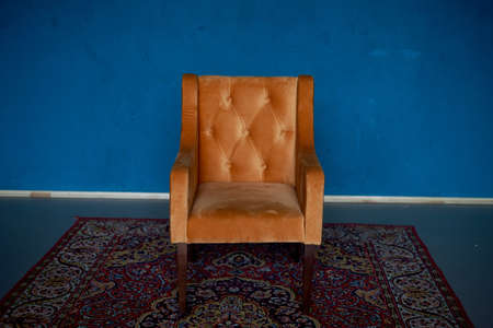 Orange retro armchair with velvet upholstery on a blue wall background. On the floor there is a red carpet with a pattern.の写真素材