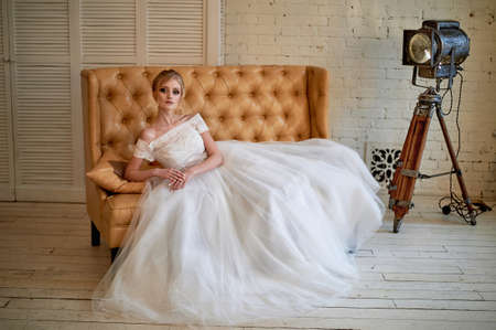 A beautiful petite blonde is sitting on the couch. The bride in a lace dress. Bride's morningの写真素材