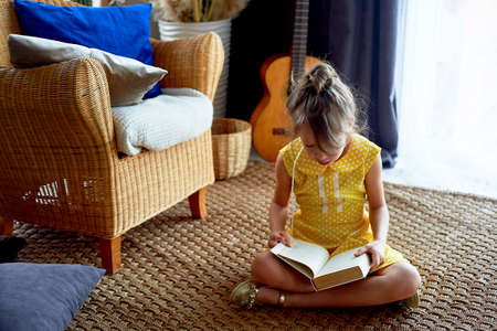 Girl, 6 years old. Her hair was pulled back in a bun. She reads the book with pleasure. Yellow dress, beautiful interior. Craving for learning, love for reading.の写真素材