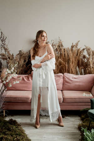 The morning of the bride. Beautiful girl in a white peniou with laces and a robe. A room with a pink sofa and cereals. aesthetics.の写真素材