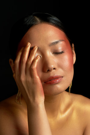 An Asian girl on a black background in the studio. Gold. Thin collarbones. The realm of beauty and perfume. Gently touches her faceの写真素材