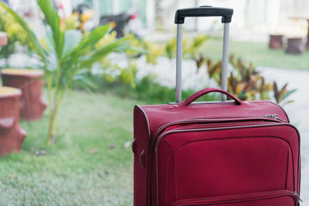 A red suitcase with a handle stands on the street against the background of the house and tropical greenery. Vacation and travel concept.の写真素材
