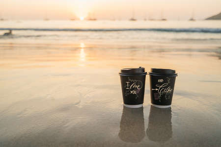 two cups of disposable black paper stand on the sand by the sea during sunsetの写真素材