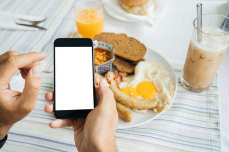 Mock up, black Smartphone in hand on a light food background. Calorie counting, weight tracking, and calorie foods. American breakfast: Fried eggs with sausages, tomatoes, bacon, and toasted bread.の写真素材