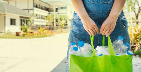 Plastic bottles are collected in a green bag in the hand of a woman against the background of a city street. Plastic collection, environmental protection.の写真素材