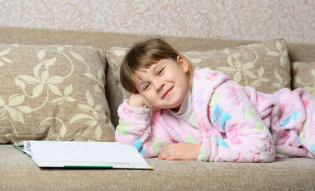 The little girl reads the book lying on a sofa. A house roomの写真素材
