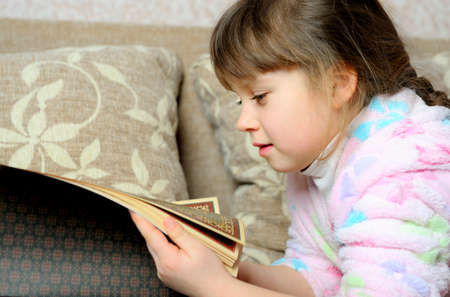 The little girl reads the book lying on a sofa. A house roomの写真素材