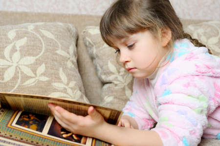 The little girl reads the book lying on a sofa. A house roomの写真素材