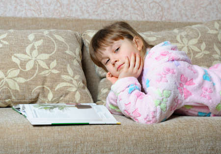 The little girl reads the book lying on a sofa. A house roomの写真素材