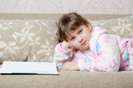 The little girl reads the book lying on a sofa. A house roomの写真素材