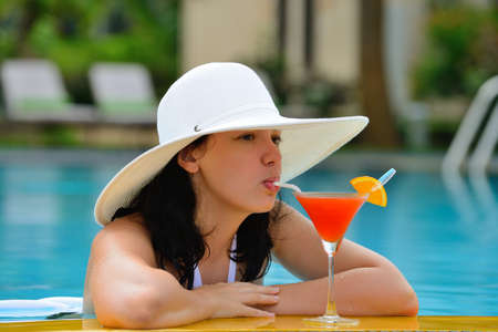 girl with a cocktail at the edge of the swimming pool . Red cocktail with an orange slice.の写真素材