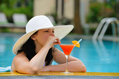 girl with a cocktail at the edge of the swimming pool . Red cocktail with an orange slice.の写真素材