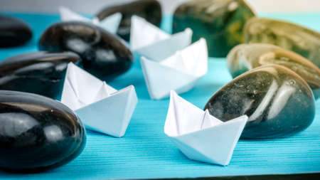 Leadership white paper boat lead further ships between abstract rock stones on blue background. Light flares right top sideの写真素材