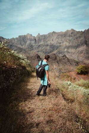 Hiker admiring barren natural landscape in front of rugged mountain range. Trekking trail on one...