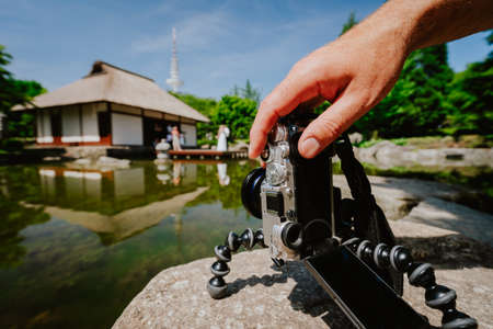 Photograph male hand handling an old vintage camera on tripod in front of water pond with landmark...