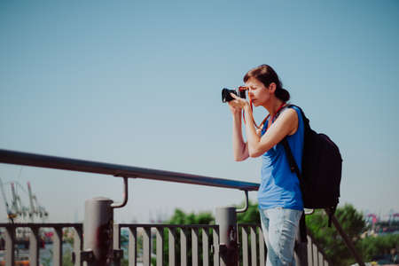 Attractive young female tourist with backpack and camera is exploring Hamburg city. Woman is making...