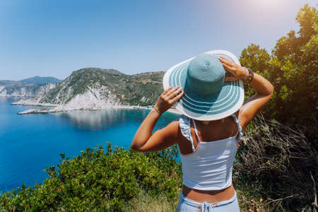 Petani beach Kefalonia. Young woman holding blue sun hat enjoying beautiful panorama of blue bay lagoon surrounded by steep cliff coastline. Greeceの写真素材