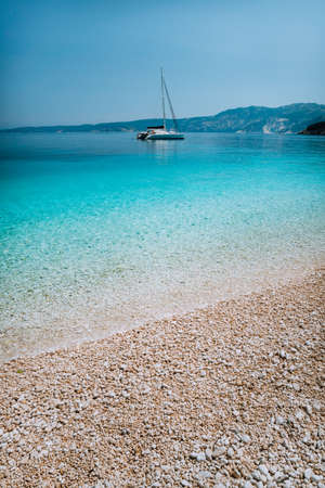 Pebble beach with pure clear azure blue water and sailing catamaran yacht boat at anchor on horizonの写真素材