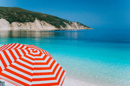 Bright red striped sun beach umbrella on beach against turquoise blue shallow sea water, white limestone coastline and sky in backgroundの写真素材