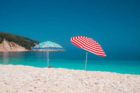 Two bright colored sun beach umbrella on pebble beach against turquoise blue sea water surface and clean blue skyの写真素材