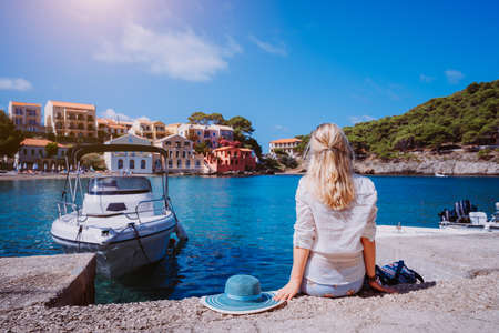 Female tourist sitting on the pier with blau sun hat laying behind. Assos village with beautiful traditional houses is in front of sea bay. Kefalonia, Greeceの写真素材