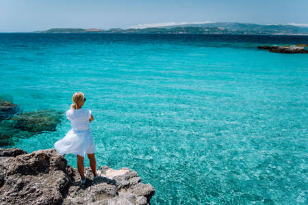 Jung adult women in white dress on summer vacation in front of sea coast landscape of small beach with crystal clear blue water. Greece, Kefaloniaの写真素材
