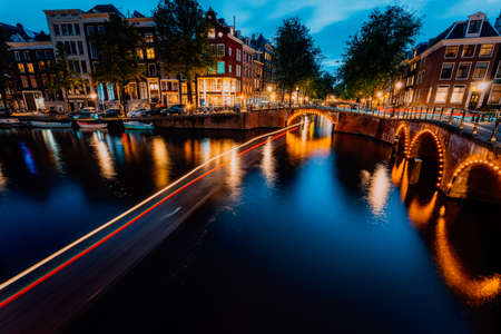 Amsterdam evening illuminated down town city. Touristic boat light trails and bridge reflections at the Leidsegracht and Keizersgracht canals. Long exposure shot. City trip visiting conceptの写真素材