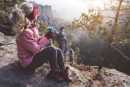 Female blond hiker outdoor clothing with coffee cup in hands on limestone rock enjoying sunset back lit view of mountain ridge and forest down the valley. Travel lifestyle adventure conceptの写真素材