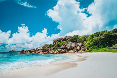 Grand Anse exotic pristine beach at La Digue island in Seychelles. White sandy beach with blue ocean lagoon, rolling waves, white clouds, granite rocks in background.の写真素材