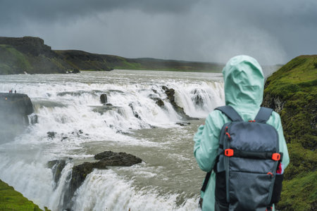 Woman with backpack and green jacket standing in front of Gullfoss powerful famous waterfall in...