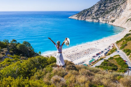 Happy woman standing on top of a rock, raising hands with an exciting feeling of freedom, looking at Myrtos Beach. Cephalonia island, Greeceの写真素材