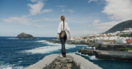 Rear view of a woman with long hair stands on the edge and raised her hands in the air in front of the ocean. Gorachico. Tenerife Islandの写真素材
