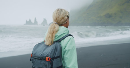 Woman with a green raincoat walking on a black sand beach enjoying view to Reynisfjara sea stack...