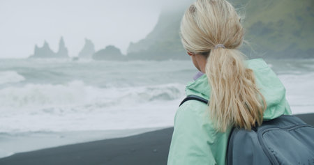 Woman with a green raincoat walking looking to silhouette of Reynisfjara sea stack cliff formations Icelandの写真素材