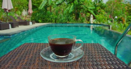Cup of coffee served by blue water swimming pool in Bali island. Green lush nature background. Weekend holiday. Outdoor cinematic static footage.の写真素材