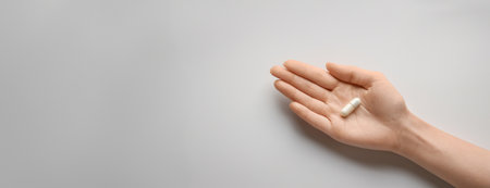 A single white capsule pill rests on the palm of a hand, displayed against a minimalist white background. Panorama with copy spaceの素材