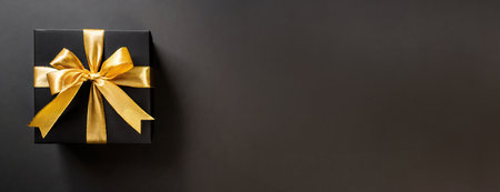 Elegant Black Gift Box with Golden Ribbon. An elegant black gift box tied with a shimmering golden ribbon sits on a dark surface, offering a luxurious and enticing presentation.の素材