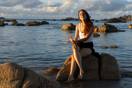 Young Galician woman on the seaside sitting on a rock at sunset near the oceanの写真素材