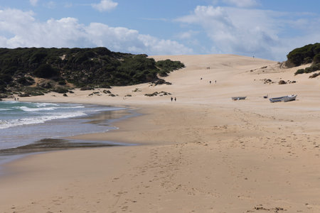 Old abandoned boat on the sand beach next to ocean with people walking on a bright sunny day with blue sky.の写真素材
